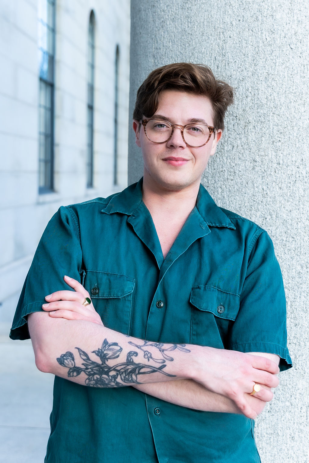 A handsome man in his mid-30s with dark hair and glasses leans against a column. His arms are crossed and his forearm tattoos are visible.